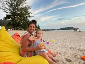 Reizigster met dochtertje zitten op een strand in Koh Lanta op een gele zitzak, met daarnaast een oranje zitzak en veel zandspeelgoed om hun heen. Links in de verte is een grote boom en rechts een eiland.