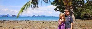 Twee jonge kinderen, links een meisje en rechts een jongen, staan tegen elkaar aan te poseren op een zandstrand in Thailand met een palmboom op de achtergrond en achter de zee een bergachtig landschap en blauwe, lichtbewolkte lucht. Het meisje steekt haar duim op.