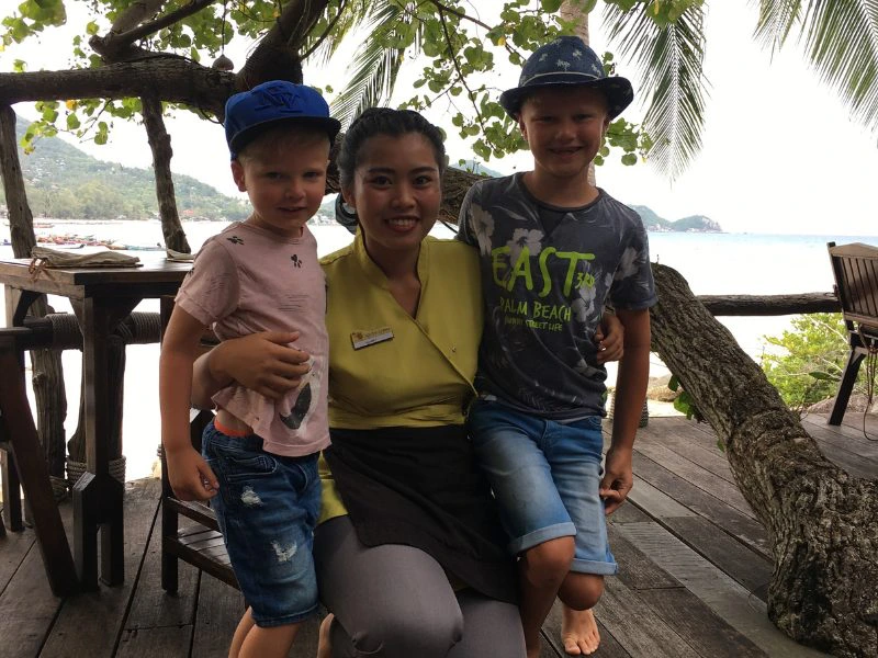 Twee jongens pet pet en hoed staan om een lokale Thaise vrouw heen die hurkt op de grond. Ze staan op een houten vlonder met een houten tafel achter hun en een schuine boom die hangt over de reling. Je ziet het strand en de zee achter hun.