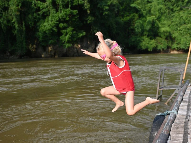 Een jong meisje springt met een druikbril en een zwemvest met haar armen omhoog de River Kwai in Thailand in. Aan het houten vlonder waar ze vanaf springt hangt ook een zwarte band. Verder is het een erg groene omgeving.