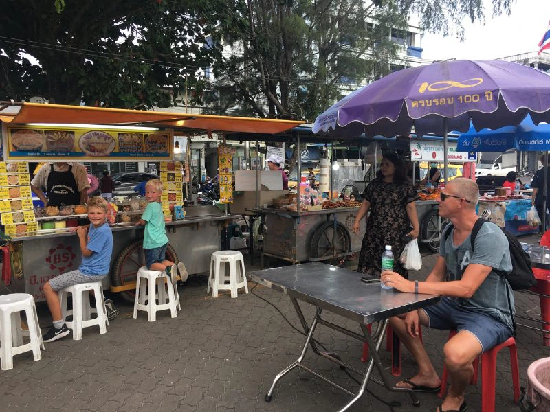 Straatscène bij een Thaise food market, met kinderen op krukjes bij een eetkraam en volwassenen aan een tafel onder een paarse parasol.