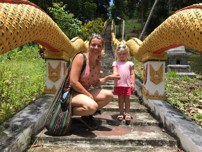 Vrouw en jong meisje poseren lachend op een tempeltrap met gouden slangenleuningen in tropische omgeving.