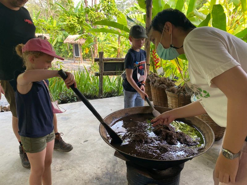Een jongen en meisje met beide een pet op staan te roeren met lange lepels in een grote pan met draadjesvlees. Hun vader staat achter hun en een Thaise man wijst naar de pan terwijl hij het meisje aankijkt. Deze man heeft een blauw mondkapje op. Het is in een groene omgeving vol palmbomen en manden met kokosnoten.