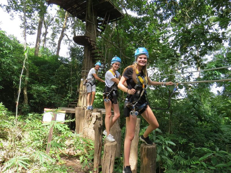 Drie mensen met helmen en klimgordels staan lachend op houten palen in de jungle, bezig met een zipline-avontuur tussen de bomen. Op de achtergrond zie je een houten platform en trappen tegen een boom.