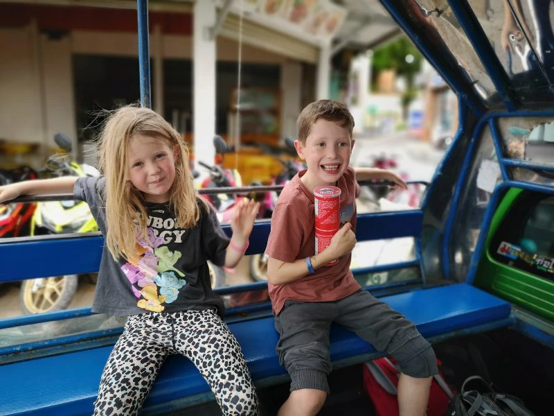 Twee jonge kinderen zitten vrolijk naast elkaar op een blauwe bank achterin een Thaise tuktuk of songthaew (openbaar vervoersbusje). Het meisje links draagt een zwart T-shirt met kleurrijke cartoonfiguren en een luipaardprint broek, en leunt nonchalant met haar arm op de rugleuning. De jongen rechts draagt een rood T-shirt en een grijze korte broek, lacht breed en houdt een rood Pringles-busje vast. Op de achtergrond zijn geparkeerde scooters en een winkelpand te zien, wat een typisch straatbeeld in Thailand geeft. De sfeer is speels en ontspannen, passend bij een familiereis.
