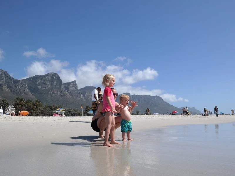 Twee jonge kinderen in badkleding staan op het natte zand aan de rand van de zee. Het meisje draagt een roze shirt en kijkt naar de golven, terwijl het jongetje in een blauwe zwembroek naast haar staat. Achter hen hurkt een volwassene gedeeltelijk zichtbaar mee. Op de achtergrond lopen strandbezoekers langs de kust en zijn hoge bergen te zien onder een blauwe lucht met witte wolken.