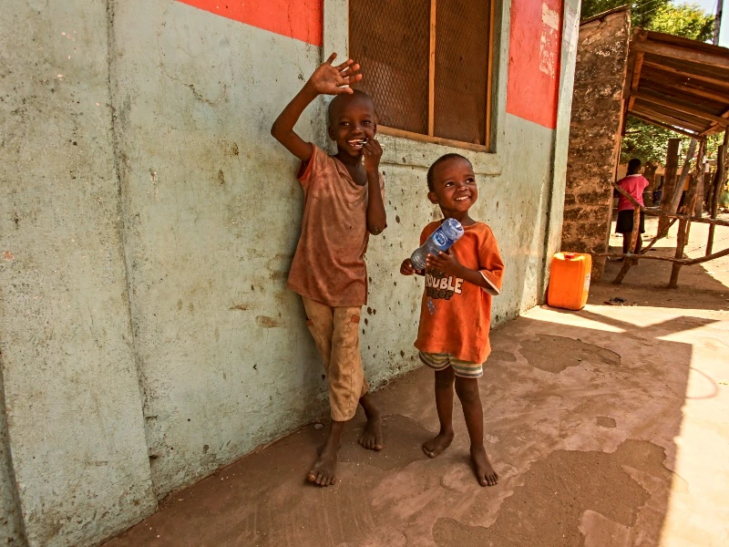 Twee jonge kinderen uit de Xhosa township staan voor een huis met rode vlakken en een houten raam. De linker jongen zwaait.