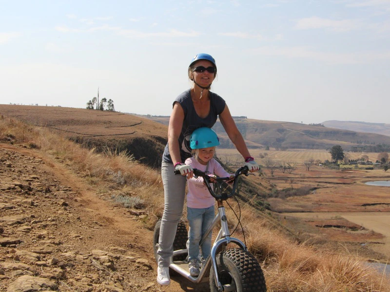 Een vrouw staat met haar dochtertje voor zich, beide met een blauwe helm op, op een step met dikke wielen in een droge omgeving met bruin zand en een blauwe lucht.