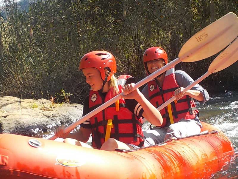 Moeder met dochter zitten in een rode raft met zwemvest aan en helm op peddelen fanatiek met achter hen bomen.