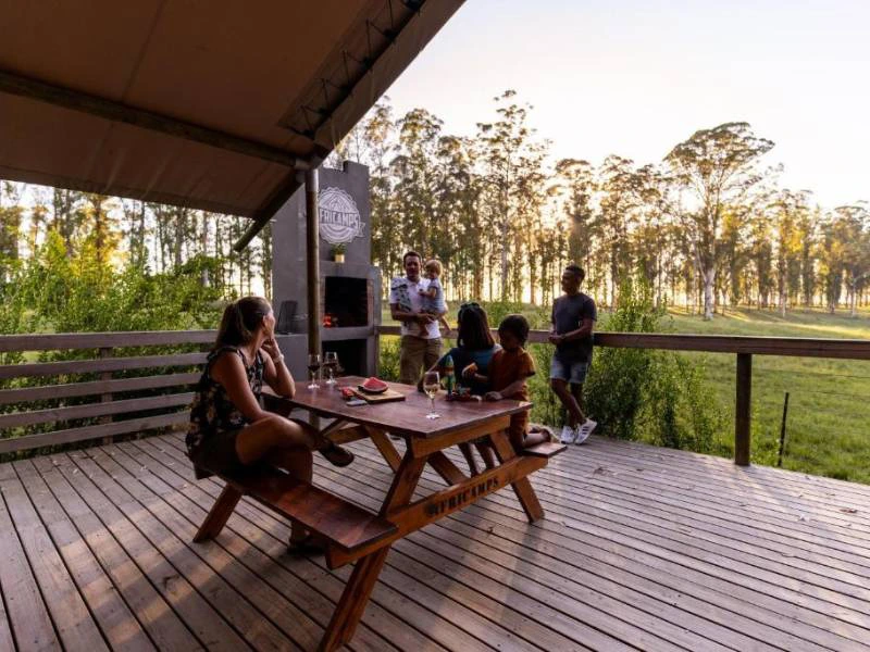 Een groep mensen geniet van een ontspannen moment op een houten terras van een glamping in de natuur van Zuid-Afrika, zittend en staand rond een picknicktafel met eten en drinken, omgeven door groene velden en hoge bomen in de avondzon.