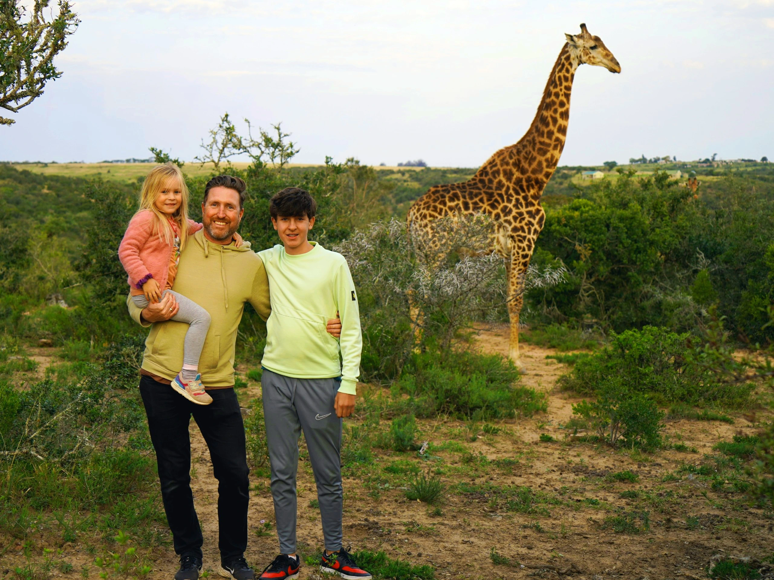 Een gezin poseert samen in de natuur tijdens een safari, met op de achtergrond een giraffe die tussen het groen staat in Zuid-Afrika.