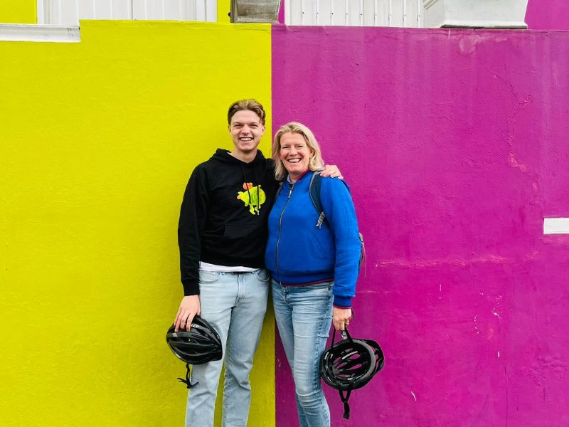 Moeder en zoon staan lachend naast elkaar voor een felgeel en knalroze muur in Kaapstad. Ze dragen casual kleding, de één in een zwarte trui, de ander in een blauwe trui, en houden beiden een zwarte fietshelm in de hand. De kleurrijke muur vormt een opvallende achtergrond die kenmerkend is voor de wijk Bo-Kaap.