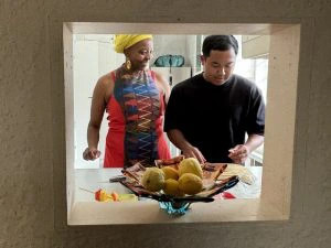 Een Zuid-Afrikaanse vrouw en reiziger staan in een doorkijkje binnen te koken. De vrouw lacht naar de man en de man is druk bezig. Voor hen staat een grote fruitschaal met fruit geel fruit erin.