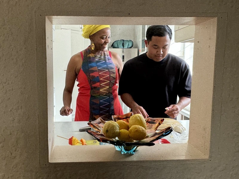 Een Zuid-Afrikaanse vrouw en reiziger staan in een doorkijkje binnen te koken. De vrouw lacht naar de man en de man is druk bezig. Voor hen staat een grote fruitschaal met fruit geel fruit erin.