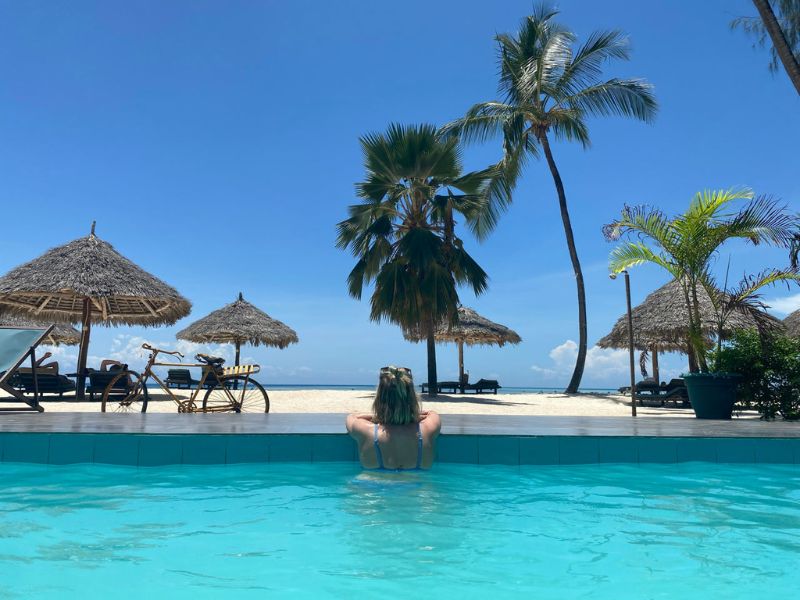 Vrouw zit in het zwembad en kijkt uit over het strand met palmbomen en rieten paraplus met in de achtergrond de zee