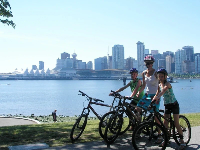 Gezin met fietsen staat naast de rivier met de skyline van Vancouver op de achtergrond.