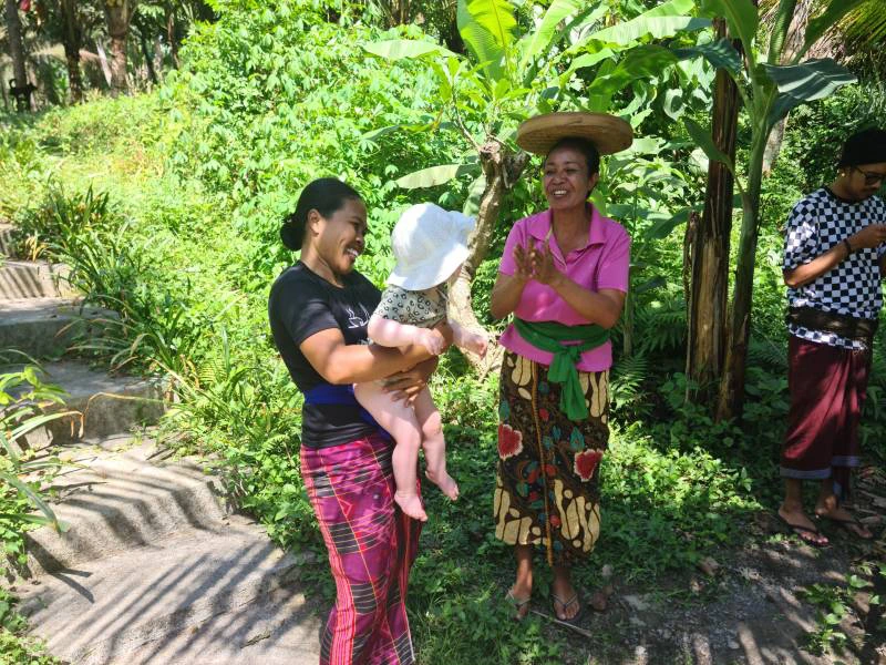 Twee Indonesische vrouwen lachen naar een klein kindje met een wit hoedje op. Een van de vrouwen heeft het kind vast. De vrouwen zijn erg kleurrijk en roze gekleed en ze staan tussen het groen.