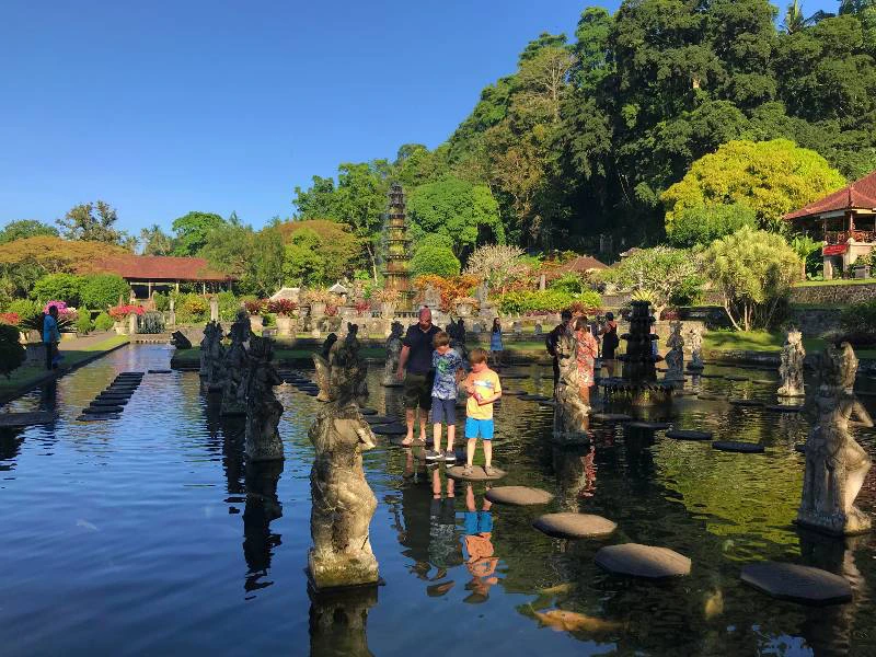 Vader met twee jonge zonen op stenen in het water bij waterpaleis op Bali. Het water weerspiegelt de stenen beelden en de groene bomen.