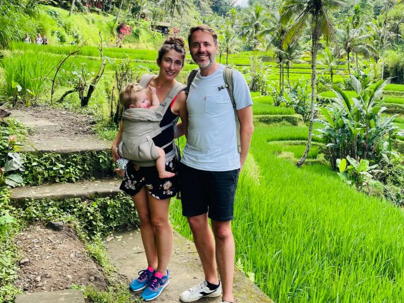 Vader en moeder met jong kind in draagzak bij moeder staan tussen de rijstvelden en palmbomen op Bali.