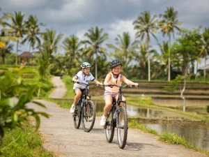 Twee kinderen die fietsen door de rijstvelden in Ubud. Er loopt een weggetje door de met water volgelopen velden en de achtergrond bestaat uit palmbomen en een donker bewolkte lucht.