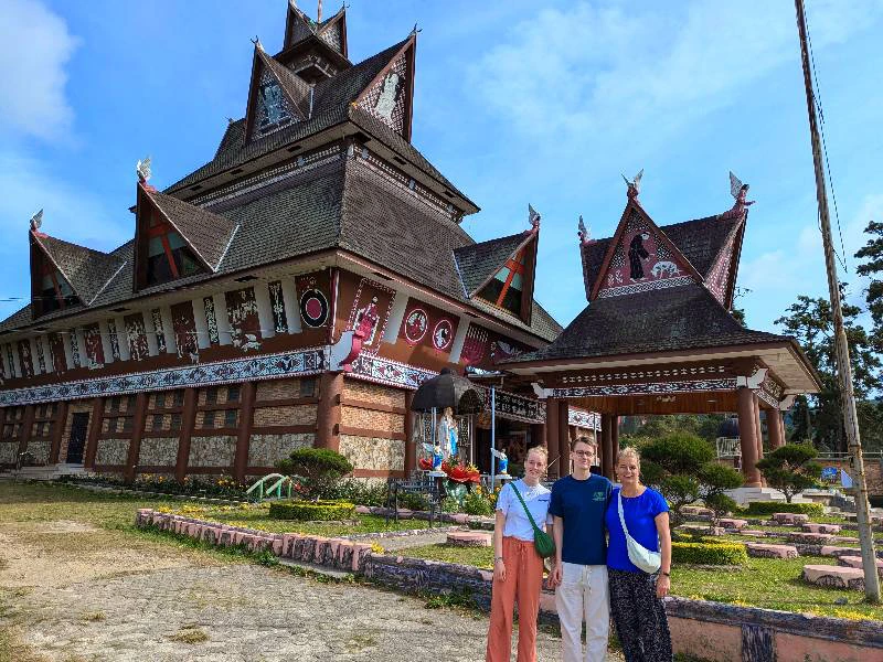 Drie mensen staan voor een traditioneel Batak-gebouw met versierde gevel op Sumatra, Indonesië.