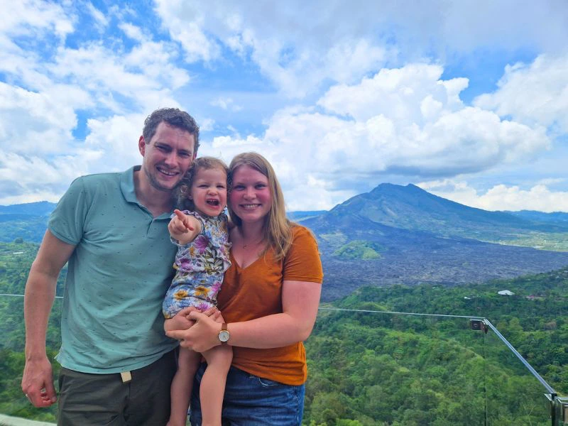 Man en vrouw houden samen een jong kind vast, met op de achtergrond het groene landschap en de vulkaan van Kintamani, Indonesië.