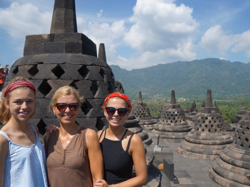 Twee tienermeiden met hun moeder in het midden lachend op de foto voor het grote tempelcomplex van de Borobudur in Java met grijze, stenen torentjes en bergen op de achtergrond. De lucht is blauw met witte wolken.