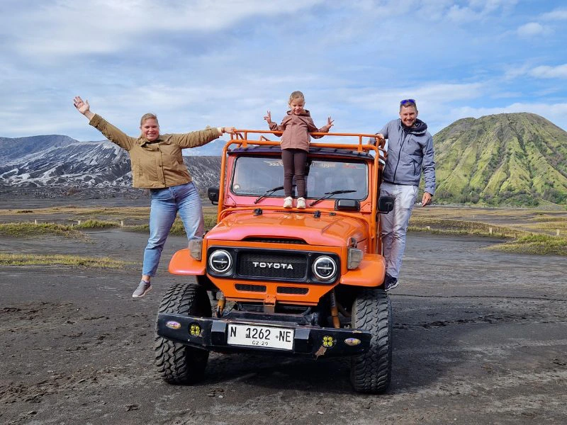 Moeder en vader hangen aan oranje jeep en dochtertje staat erop. De ondergrond is modderig en achter het groene bergachtige landschap te zien van de Bromo in Java.