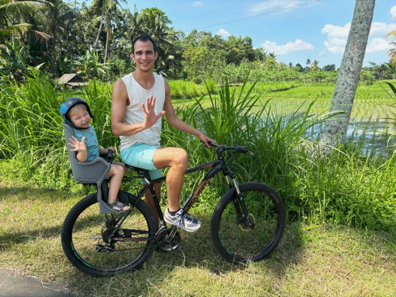 Vader staat stil op een zwarte fiets met zoontje achterop met een blauw helmpje op. Ze zwaaien en lachen allebei. Ze staan stil in het gras en achter hun zie je de rijstvelden iets lager geleden.