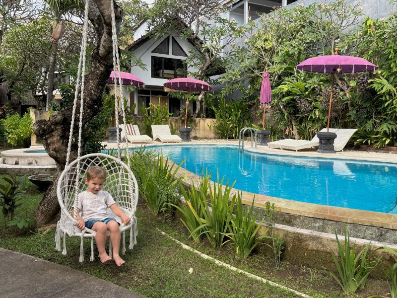 Een jongetje zit in een wit gevlochten stoel die hangt aan een boom naast het zwembad in een hotel in Amed. De omgeving is groen en er liggen ligbedden met roze parasollen.
