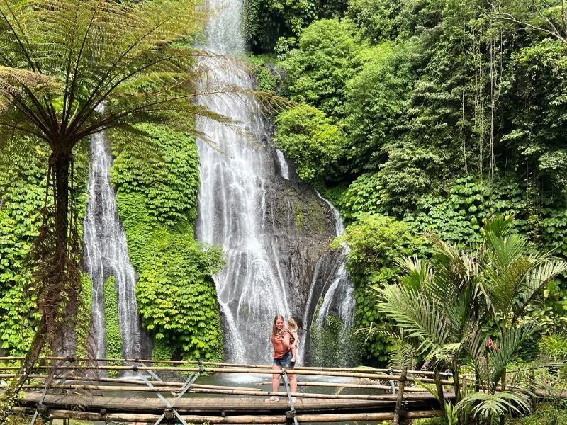 Moeder met dochtertje op haar rug staat op een houten brug met achter haar een hoge waterval en veel groen om haar heen van struiken en palmbomen.
