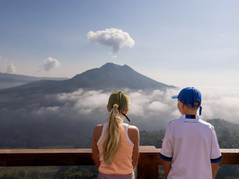 Een meisje met een staart in en een jongen met een pet op vanaf de achterkant kijken naar de Mount Batur met een paar witte wolken eromheen en een blauwe lucht.