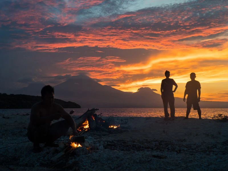 Een roze, oranje, gele lucht van de ondergaande zon met wolken en bergen in de verte. Op de voorgrond is een strand te zien met iemand links in beeld die een kampvuur aansteekt en rechts staan twee mensen ernaar te kijken.