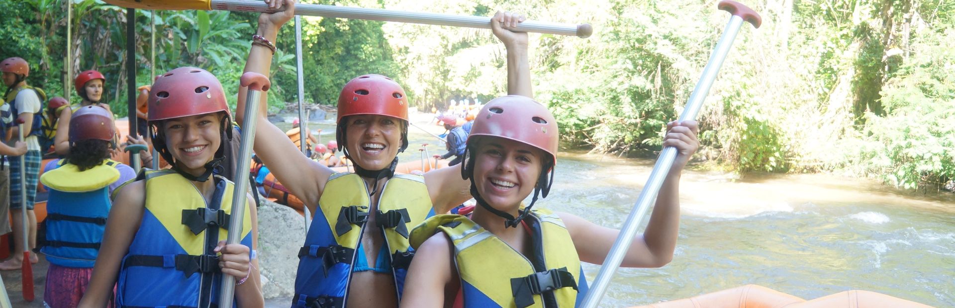 Moeder met naast haar aan beide kanten haar dochters. Alle drie erg blij en lachend in de camera met geel/blauwe zwemvesten aan ze houden hun peddels omhoog. Achter hun zie je de rivier en veel groen.