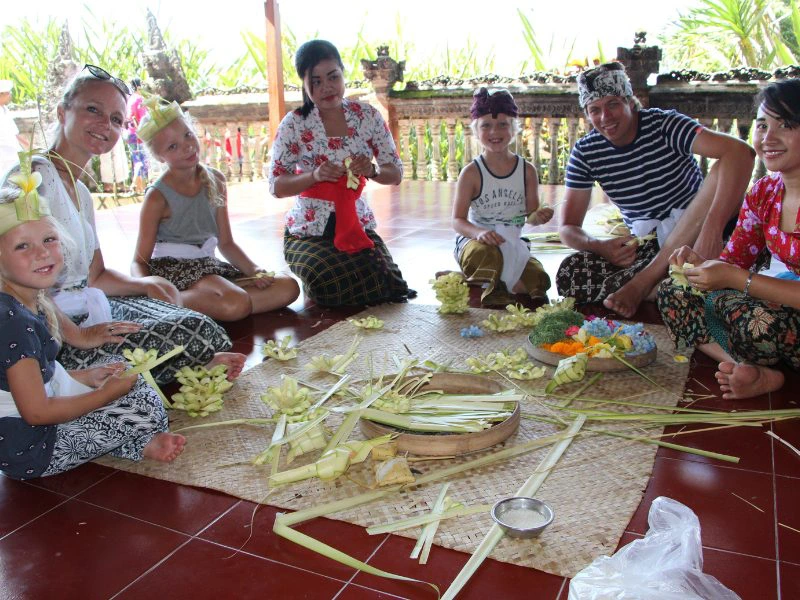 Groep van zes mensen (drie kinderen en drie volwassenen) zit in een kring op een bamboemat terwijl ze traditionele Balinese offers maken van palmblad. Op de achtergrond een stenen muurtje met uitzicht op groene natuur. Iedereen draagt traditionele kleding of een sarong en lacht naar de camera.