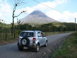 Een zilverkleurige auto rijdt over een onverharde weg met op de achtergrond de Arenal-vulkaan in Costa Rica.