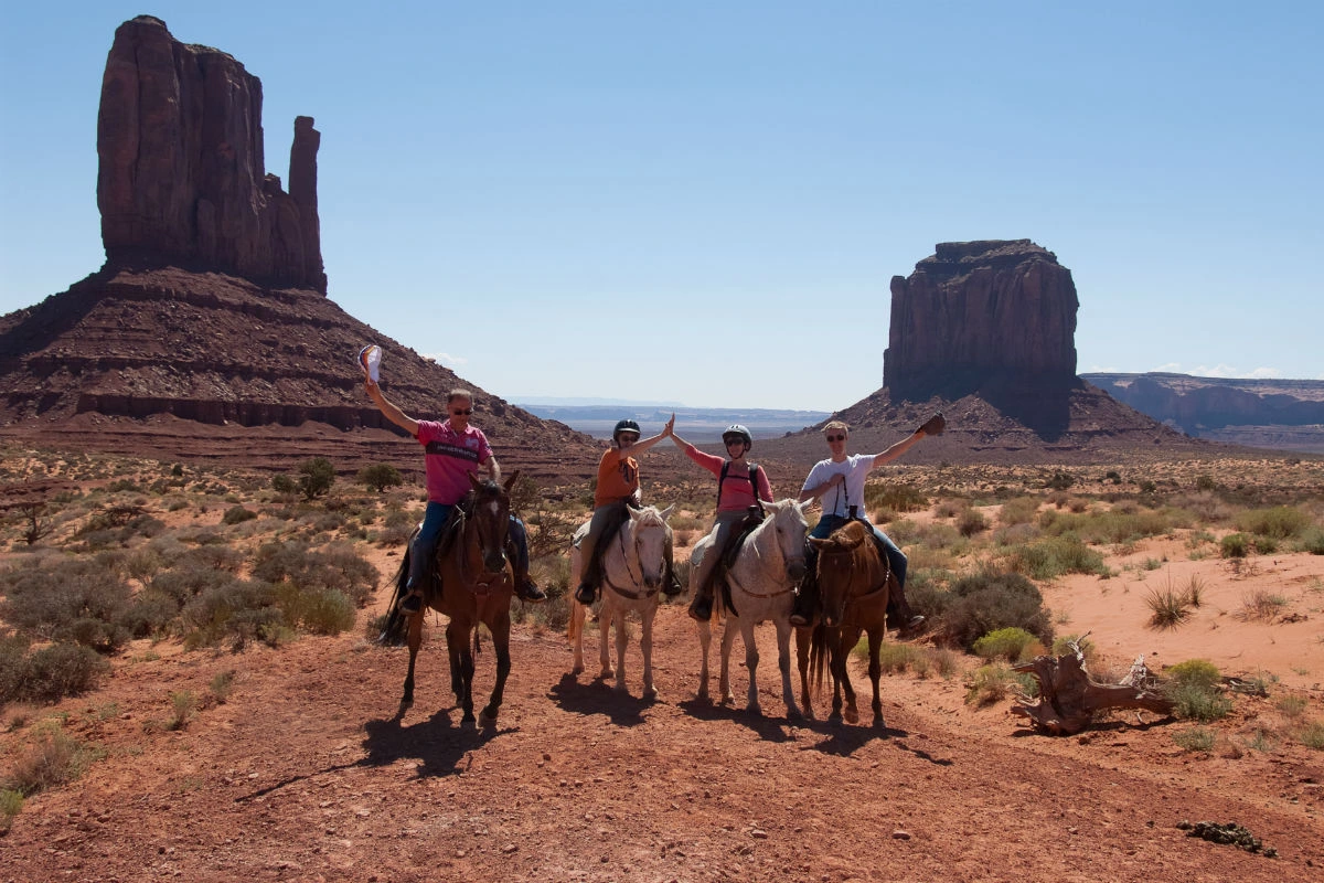 Vier mensen te paard in Monument Valley, met de iconische rode rotsformaties en een blauwe lucht op de achtergrond.