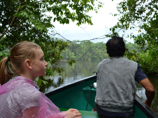 Meisje in een roze poncho en een gids varen in een groene boot door een rustige rivier, omgeven door dichte jungle.