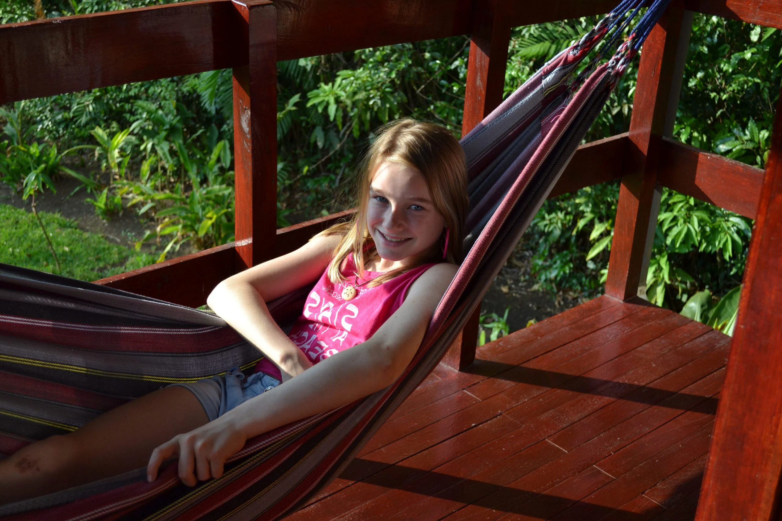 Meisje ligt ontspannen in een kleurrijke hangmat op een houten veranda, omringd door groene tropische planten.
