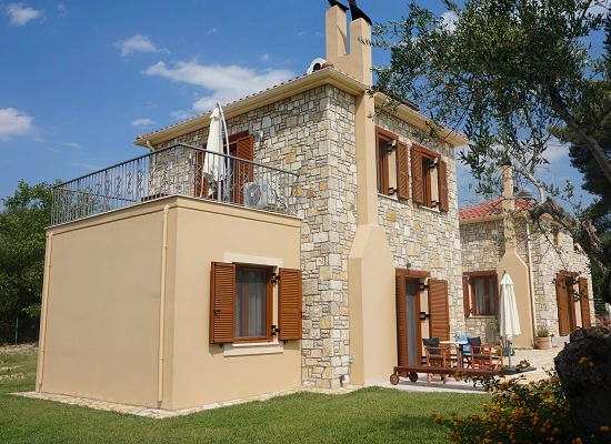 Twee verdiepingen tellend huis van natuursteen met houten luiken en terras, omringd door gras en bomen.