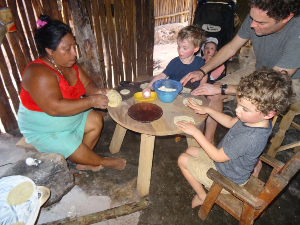 Familie maakt samen met een lokale vrouw tortilla’s aan een houten tafel in een eenvoudig huisje.