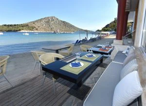 Gedekte eettafels met uitzicht op een strand en rustige baai met heuvels en bootjes op het water.