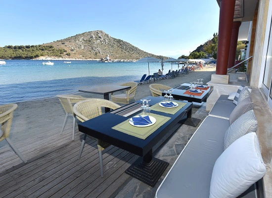 Gedekte eettafels met uitzicht op een strand en rustige baai met heuvels en bootjes op het water.