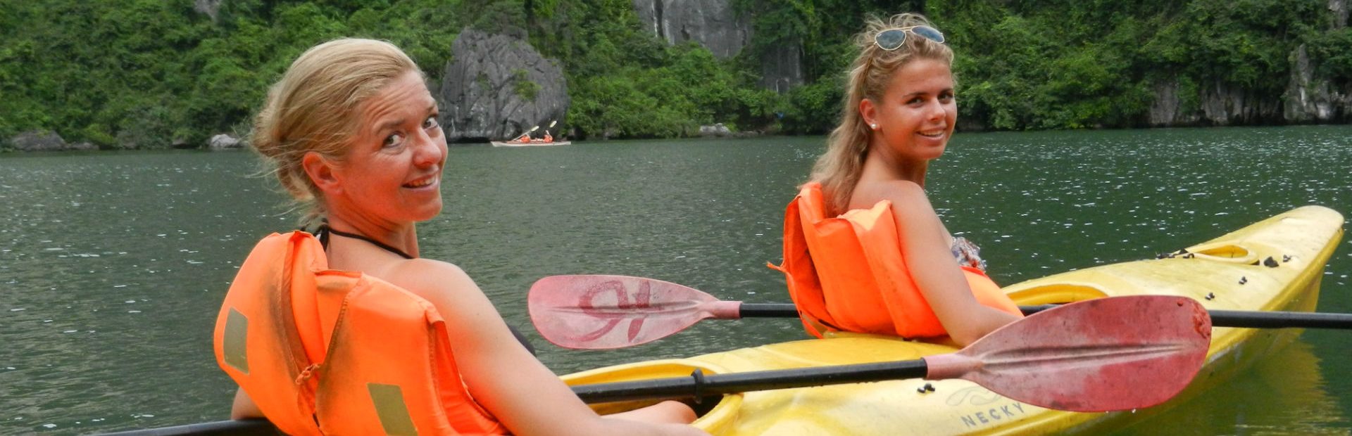 Twee vrouwen in een gele kajak met oranje reddingsvesten, peddelend door een groene baai omringd door rotsen en jungle.