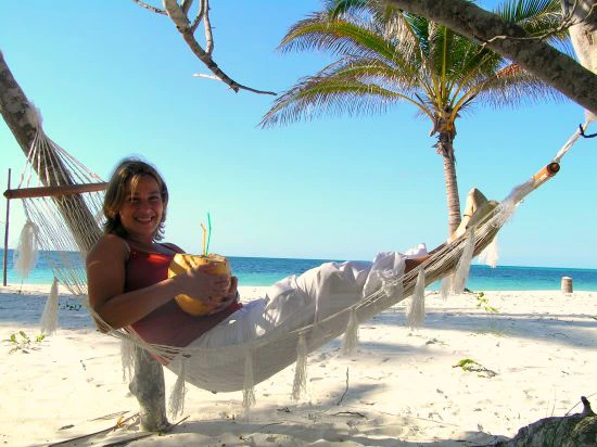 Vrouw ligt ontspannen in een hangmat op een tropisch strand met een kokosnootdrankje in haar hand, onder een palmboom met uitzicht op de zee.