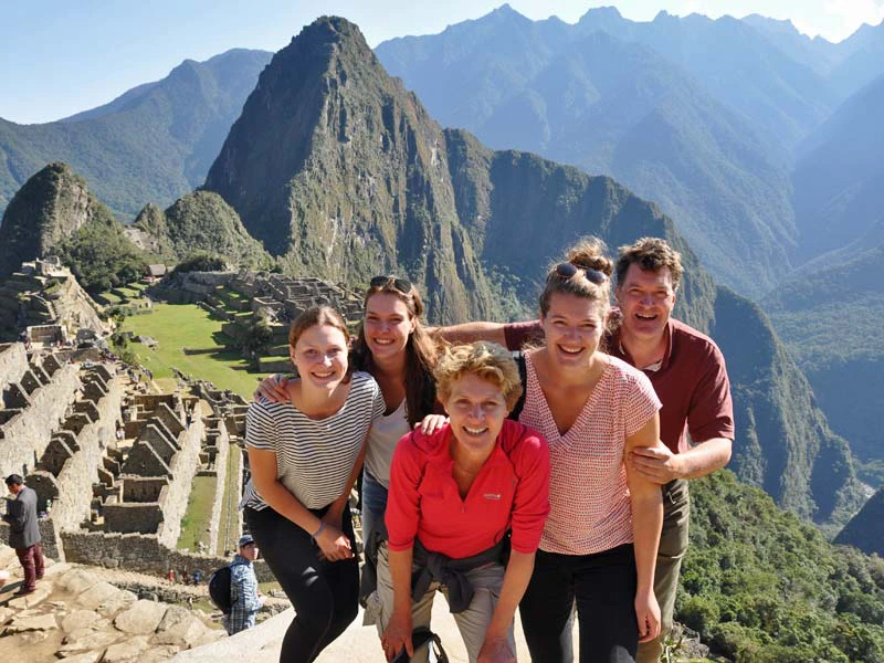 Groep van vijf mensen poseert lachend bij Machu Picchu met groene bergen op de achtergrond.