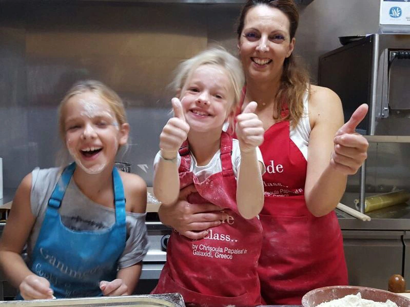 Moeder en twee jonge dochters in een keuken dragen schorten en lachen, terwijl ze met deeg bezig zijn en duimen omhoog steken.