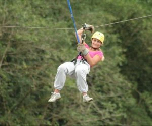 Vrouw met helm en veiligheidstuigje glijdt lachend aan een zipline door een groen bosrijk gebied.