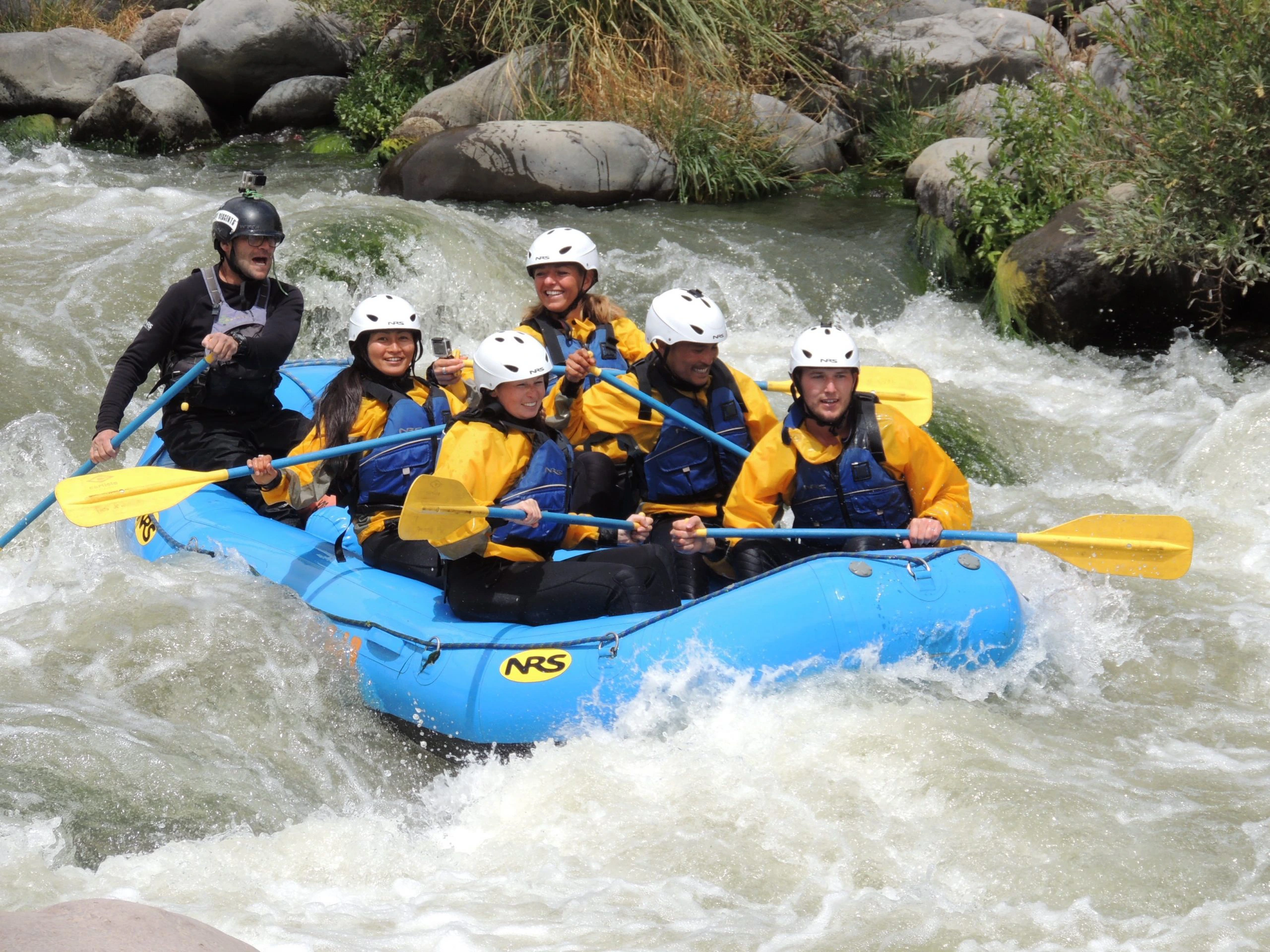 Groep mensen in een blauwe raft met helmen en peddels vaart door wilde stroomversnellingen tijdens een raftingtocht op een rivier.