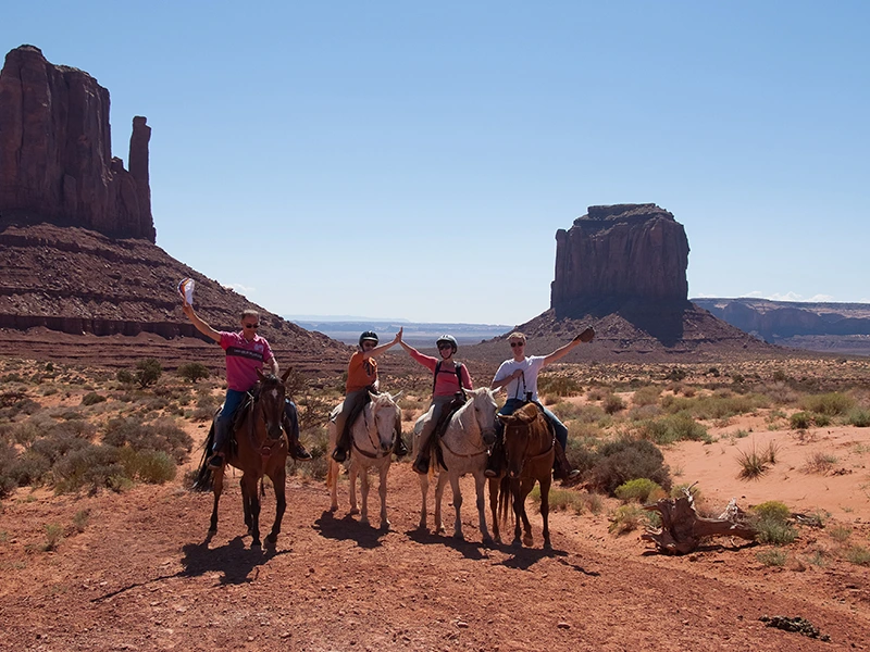 Vier mensen te paard in Monument Valley, met de iconische rode rotsformaties en een blauwe lucht op de achtergrond.